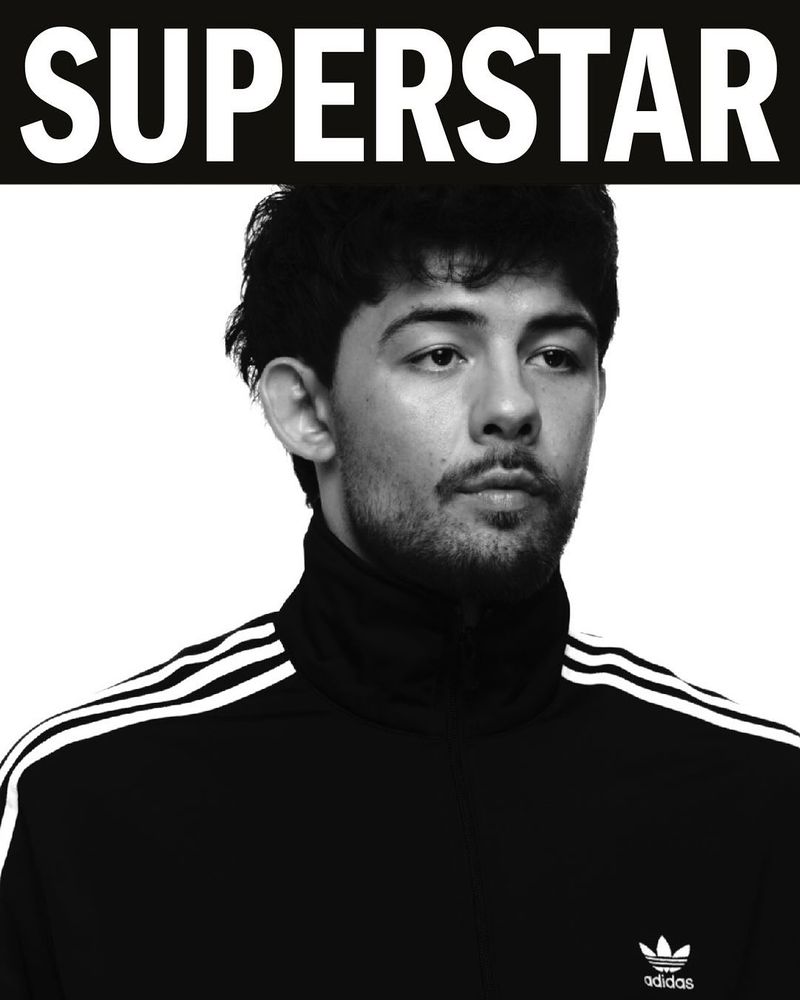 Portrait of a male with short, dark hair and a slight beard, wearing a black Adidas tracksuit jacket, against a white background.
