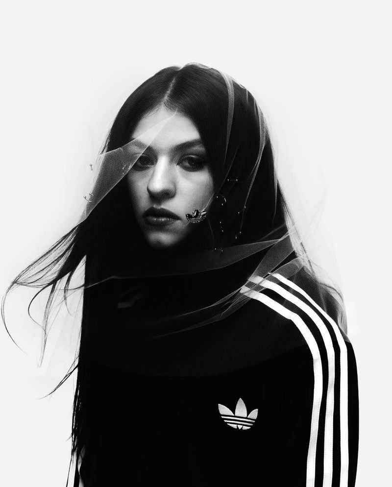 Monochrome portrait of a woman with long dark hair and a translucent veil, wearing an Adidas track jacket with white stripes.