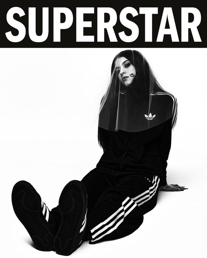 Black and white fashion shot of a woman wearing Adidas tracksuit and veil while sitting against a white background.