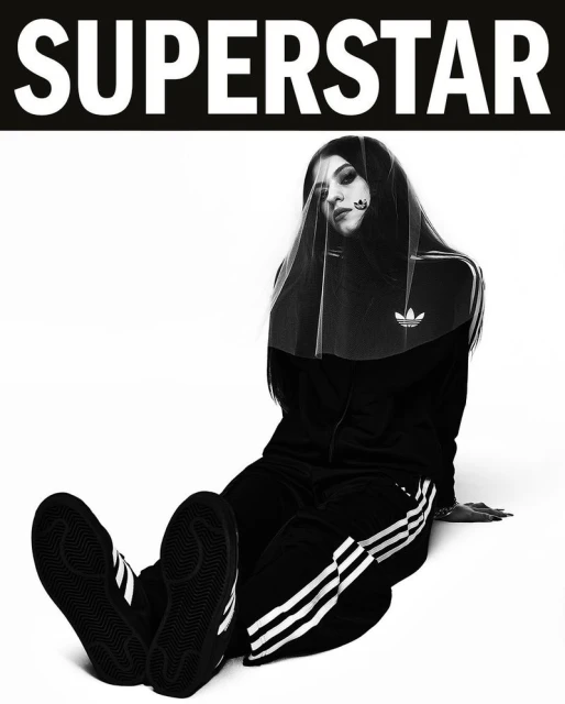 Black and white fashion shot of a woman wearing Adidas tracksuit and veil while sitting against a white background.