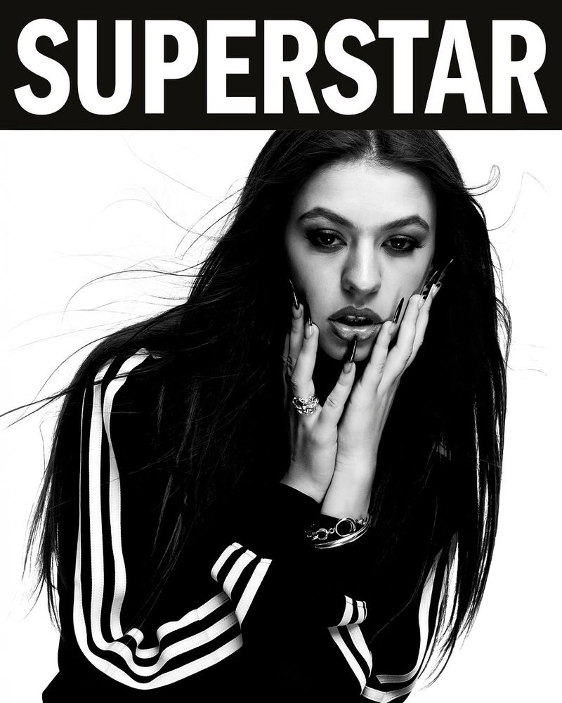 Portrait of a young woman with long dark hair and dark eye makeup, wearing a striped jacket and jewelry, with the text SUPERSTAR above.