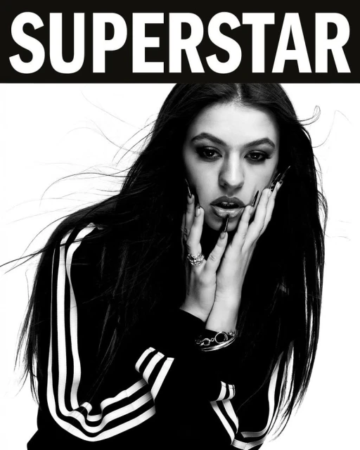 Portrait of a young woman with long dark hair and dark eye makeup, wearing a striped jacket and jewelry, with the text SUPERSTAR above.
