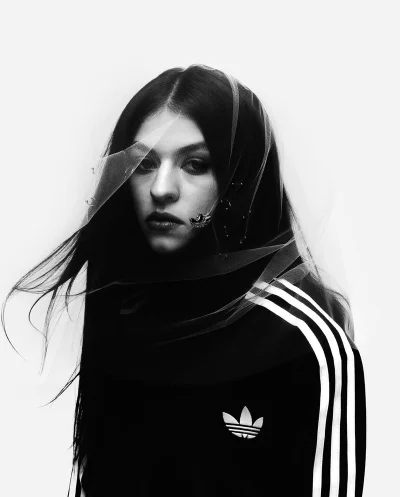 Portrait of a woman wearing a black Adidas track jacket and a sheer veil against a white background.