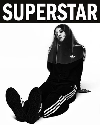A woman seated on a white floor wearing a black Adidas tracksuit with white stripes, black sneakers, and a sheer veil over her head.