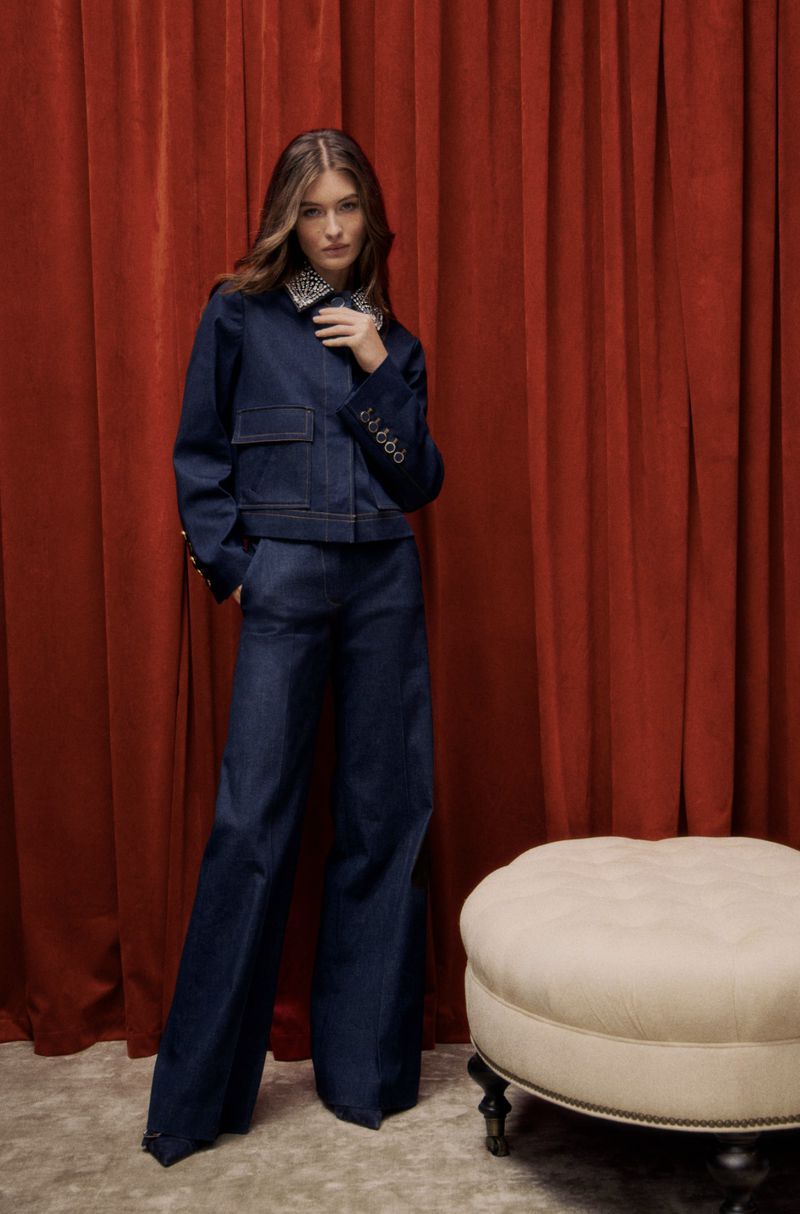 A young woman in a blue denim outfit strikes a pose in front of a red curtain, with an ottoman visible in the lower right corner.