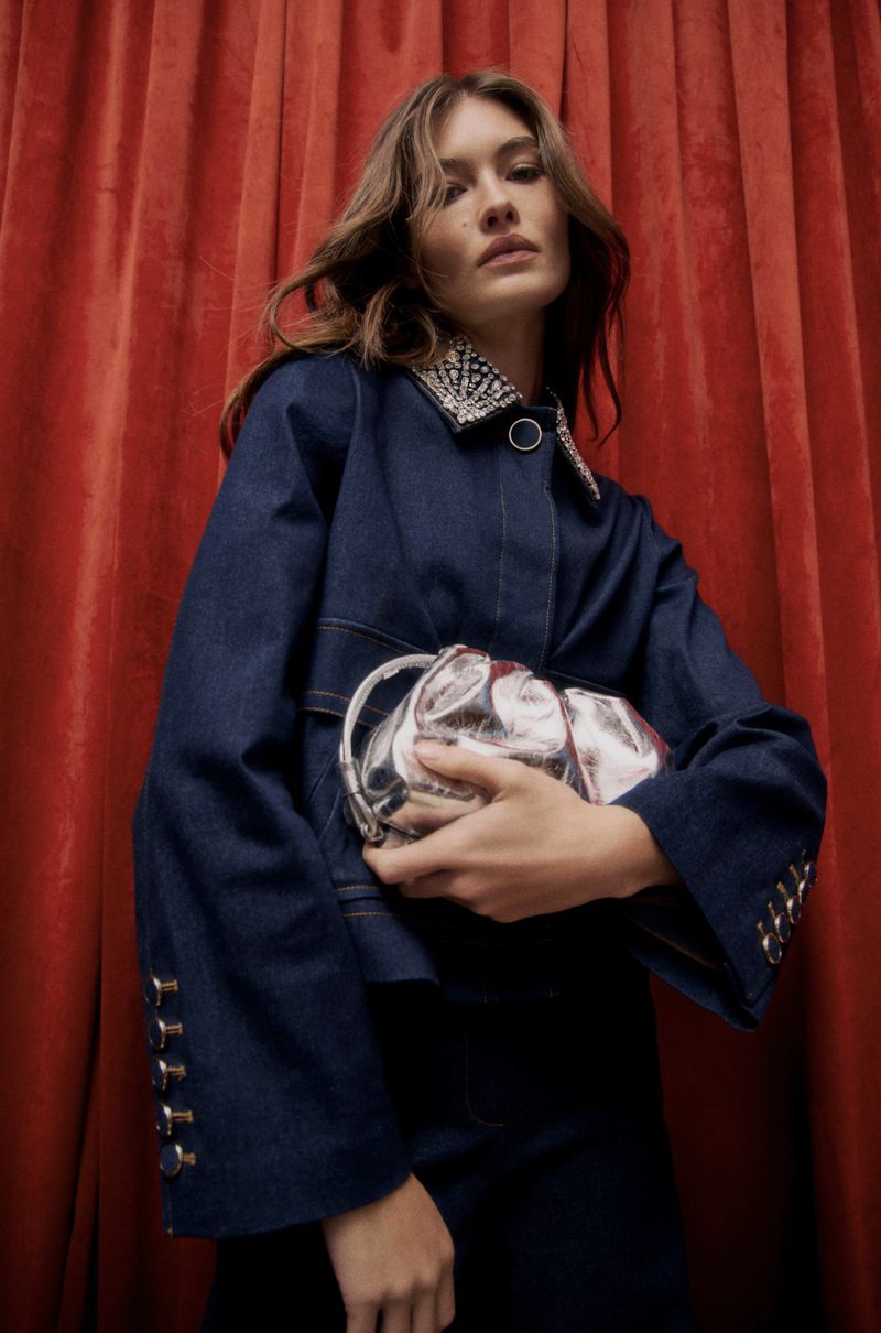 A fashionable adult female with brunette hair is holding a silver clutch bag in a low-angle shot against a red curtain backdrop.