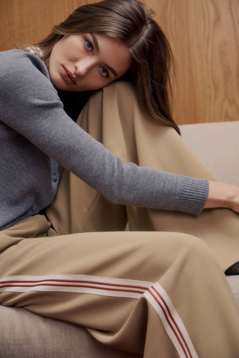 Attractive brunette woman gazes at the camera while resting her head on her arm, wearing a gray sweater and beige pants with side stripes.