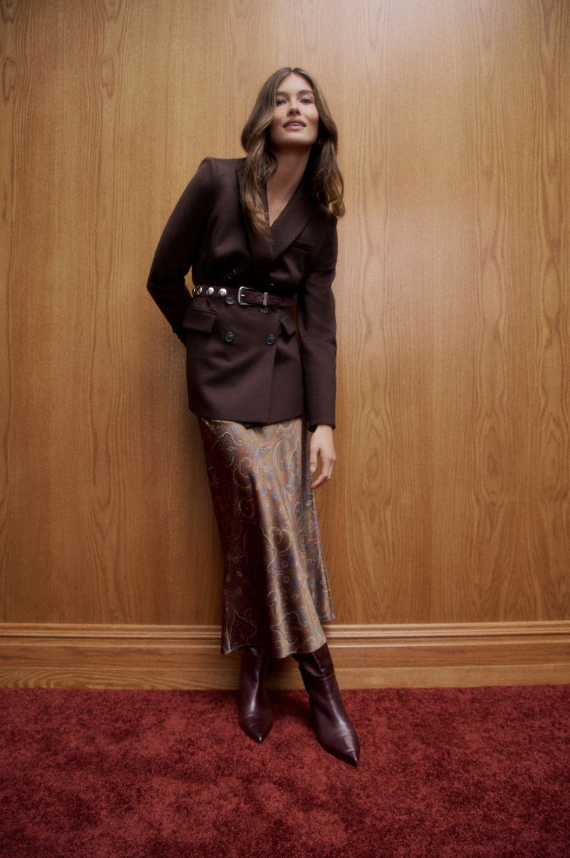 Elegant woman poses in brown blazer, paisley skirt, and matching boots against a wood-paneled wall and burgundy carpet.