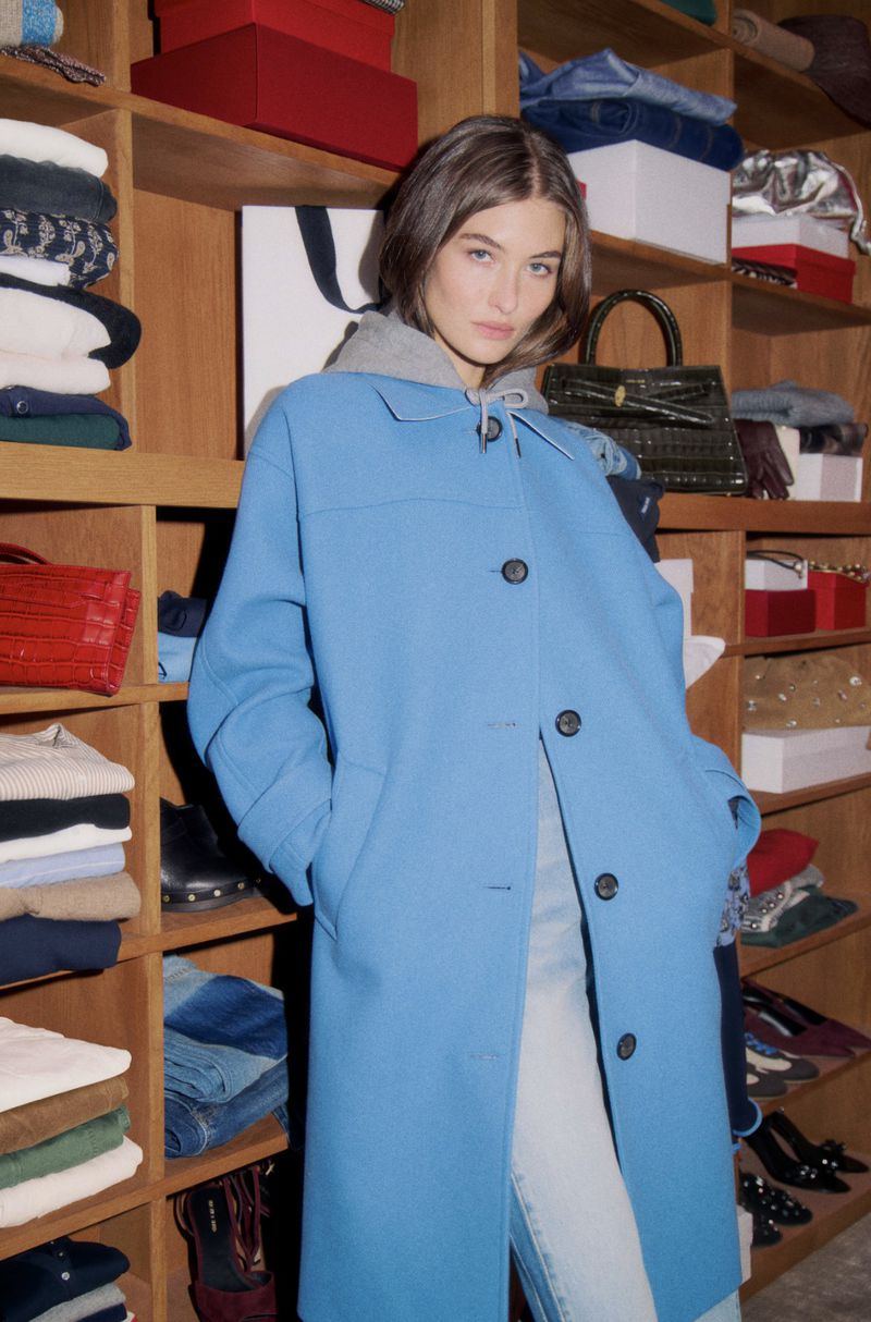 A woman poses indoors, wearing a blue coat and jeans, next to shelves displaying clothing and accessories in a well-lit environment.