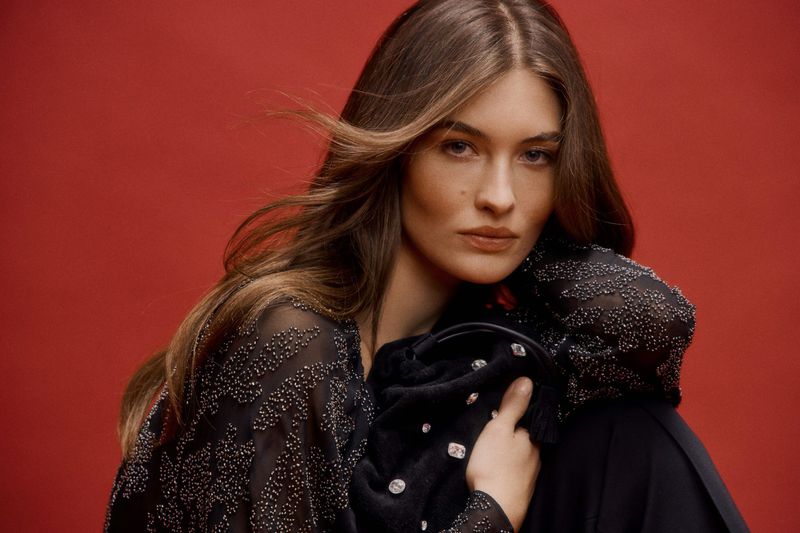 Woman in embellished sheer top poses with a jeweled handbag against a red backdrop in a studio setting, showcasing fashion and beauty.