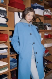Model Grace Elizabeth wearing a bright blue buttoned coat over a grey hoodie in front of wooden wardrobe shelves.
