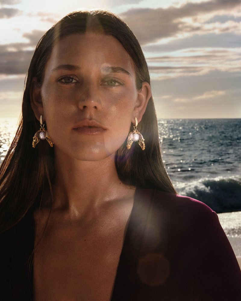 Vivienne Rohner wearing ornate gold and pearl drop earrings against a backdrop of ocean waves and a clouded sunset sky.