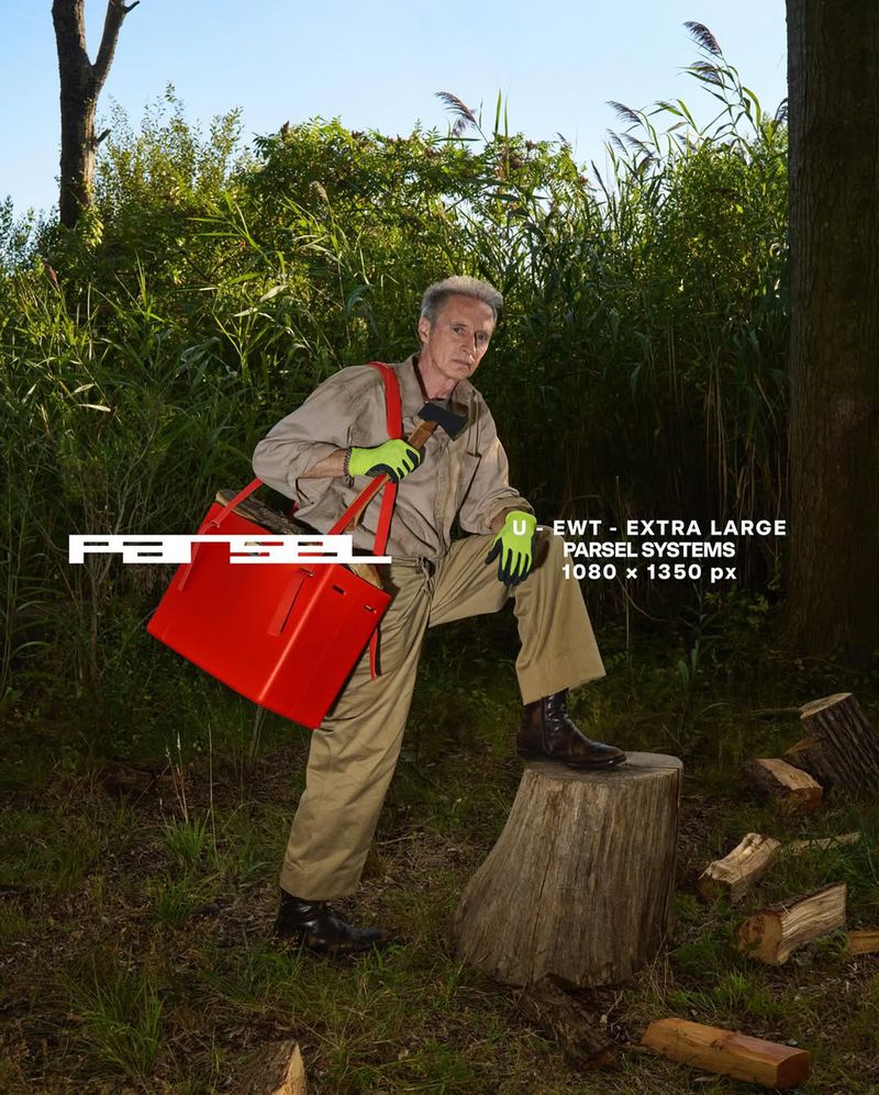 Adult male posing outdoors in a forest. He is wearing work clothes and holding a red bag with a tree stump next to him.