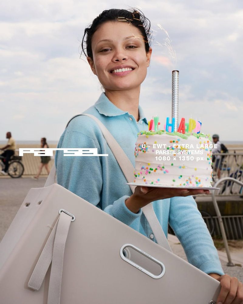 Woman smiles, holding a birthday cake outdoors near a beach. She wears a light blue shirt and carries a beige bag.