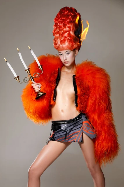 Stylish woman in a fur coat and skirt poses with a candelabra, featuring elaborate hair with a burning flame effect, against a grey background.