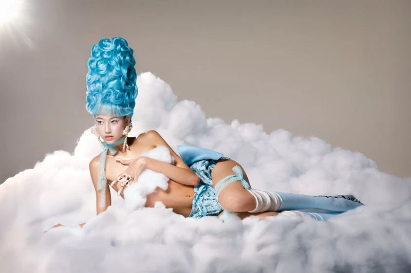 Adult female model wearing a blue Marie Antoinette style wig and partial costume, posing in a cloud-filled indoor environment with studio lighting.