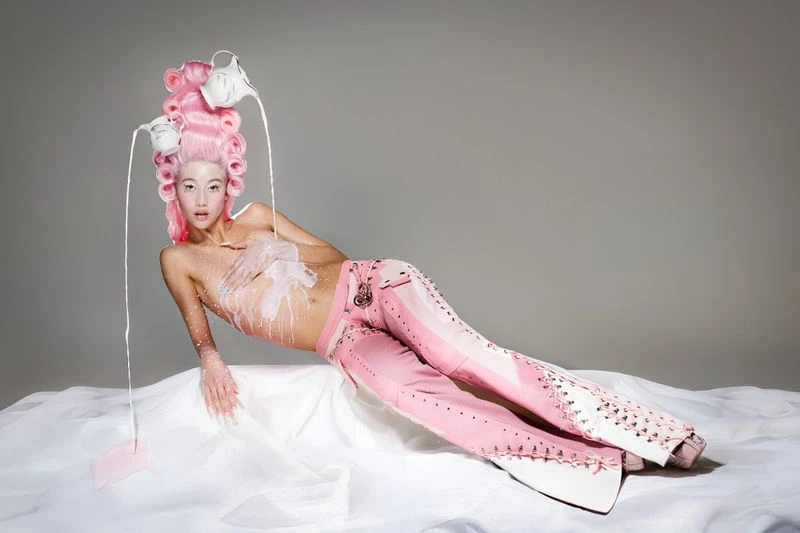 Portrait of a woman with pink wig and pink pants, lying on a white sheet with white liquid running over her in a studio setting.