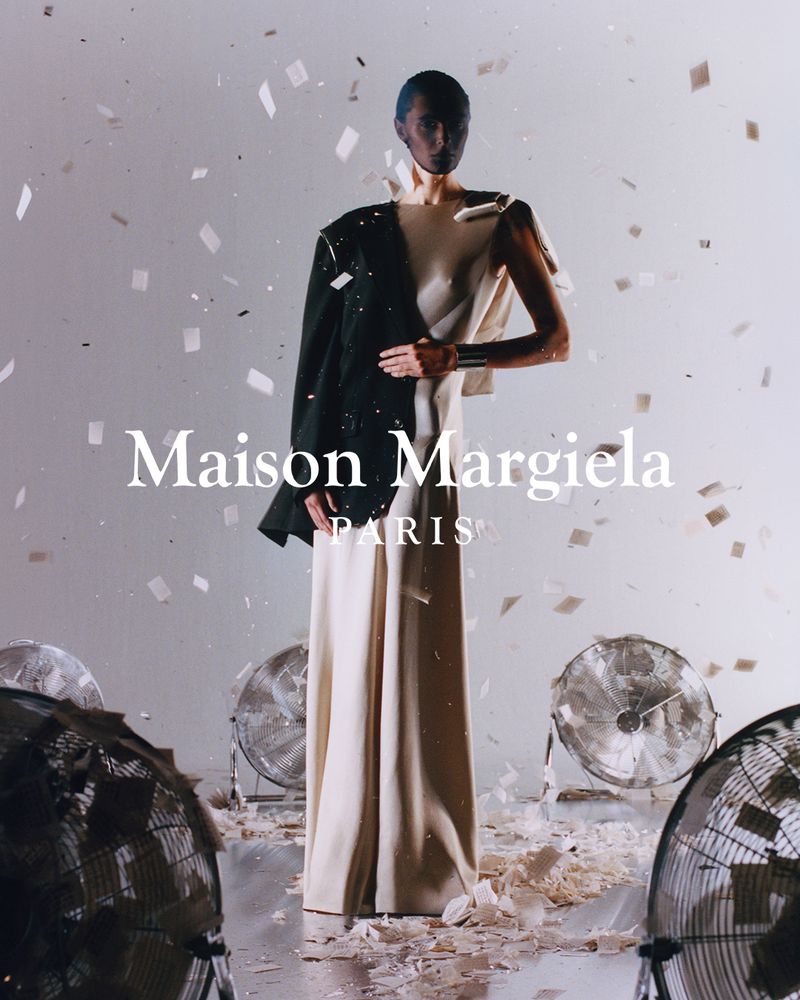 A fashion shot featuring a slender person in a long cream dress with a dark jacket, confetti, fans, and the Maison Margiela logo.