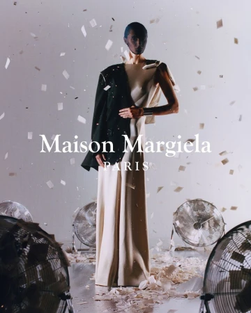 A fashion shot featuring a slender person in a long cream dress with a dark jacket, confetti, fans, and the Maison Margiela logo.