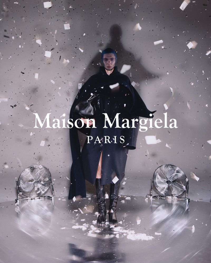 Fashion shot of a model in a dark cape-like dress with knee-high boots, standing among confetti and fans on a reflective grey surface.