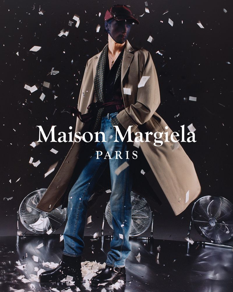 A male fashion model in a tan overcoat stands in front of fans, under confetti, showcasing Maison Margiela Paris apparel and accessories.