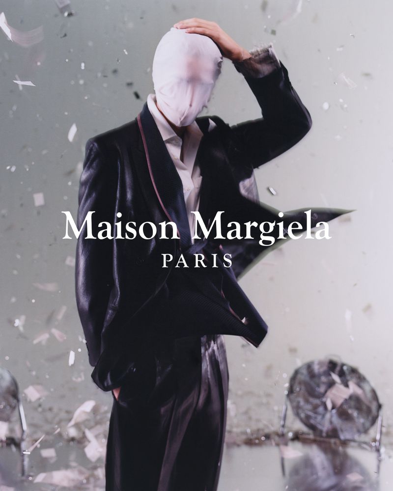 Fashion model in a black suit with a white head covering posing in a studio with confetti. Maison Margiela PARIS text overlay.