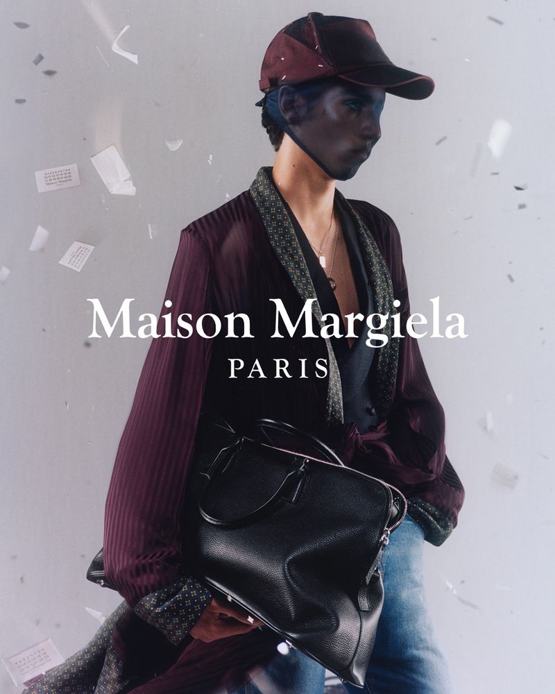 Fashion shot of a model in a cap, robe, and jeans, holding a black duffel bag with the Maison Margiela logo visible against a white background.