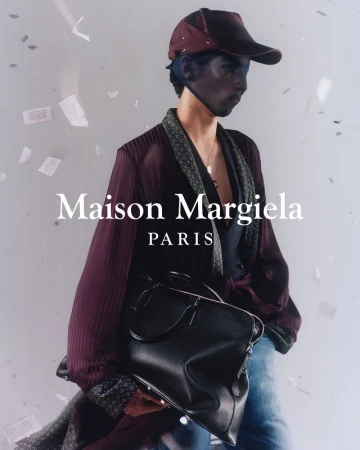 Fashion shot of a model in a cap, robe, and jeans, holding a black duffel bag with the Maison Margiela logo visible against a white background.