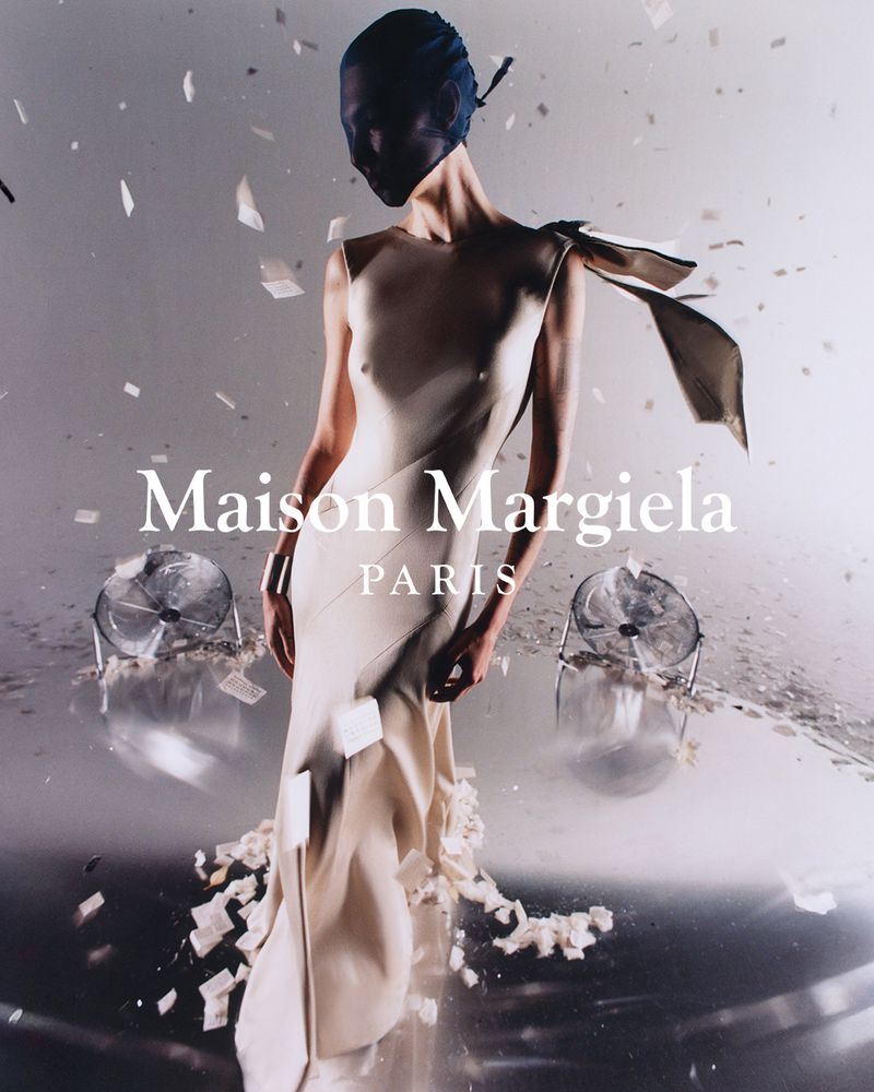 Fashion model in a flowing white gown with artistic draped detail, wearing a dark head covering, posing in a surreal, light-filled space.