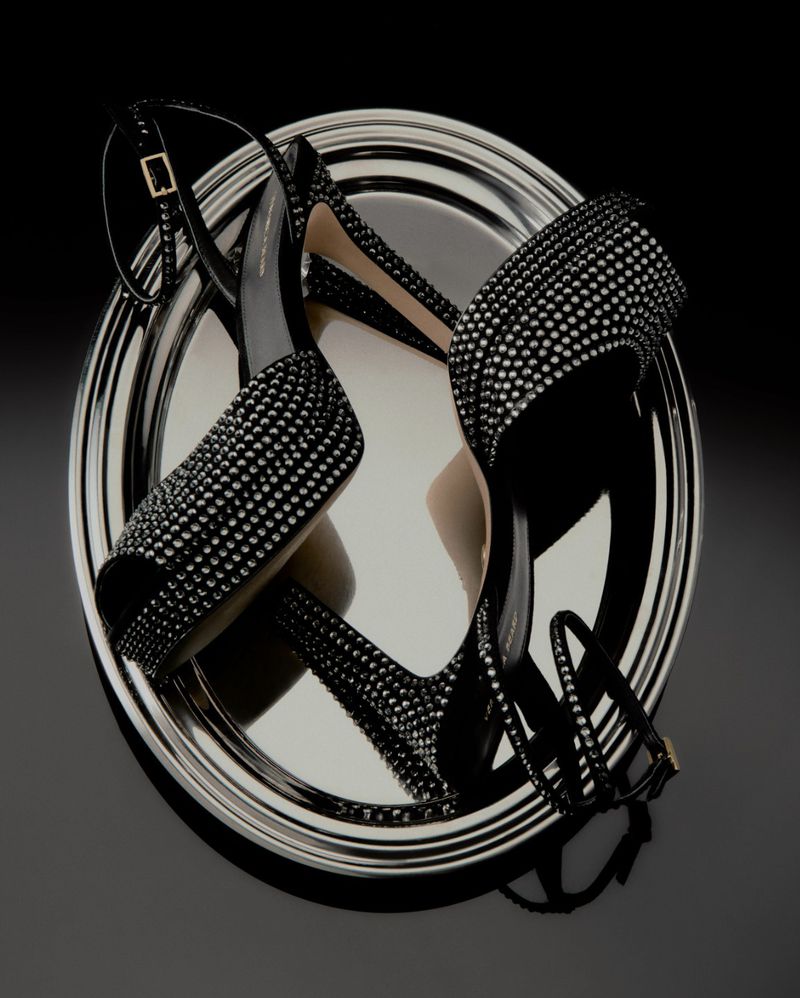 High-heeled sandals with decorative studs displayed on a silver tray, set against a dark background, showcasing fashion footwear.