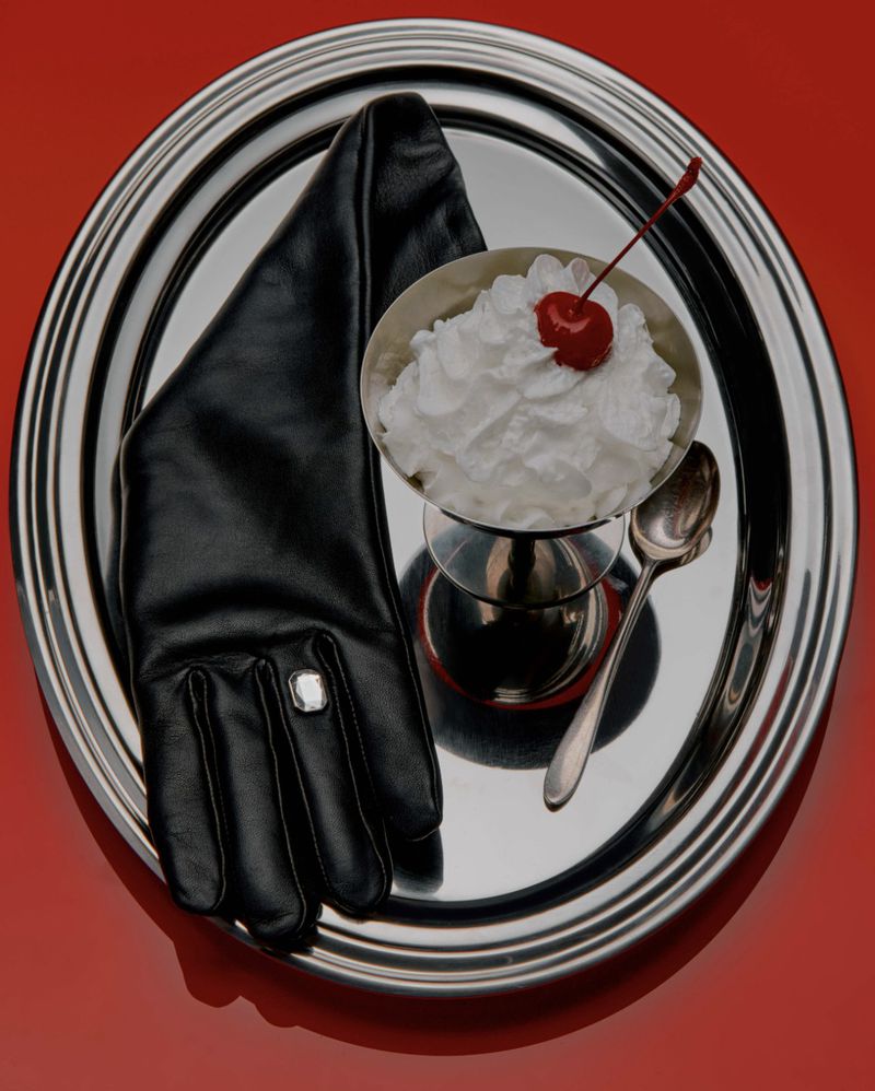 Overhead shot of a silver tray with a black leather glove, a dessert cup with whipped cream and cherry, and a spoon on a red background.