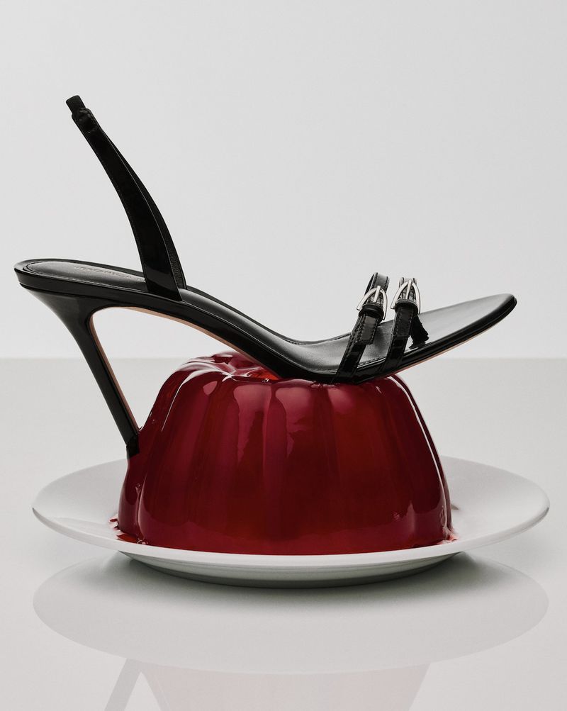 Black high-heeled sandal on a red gelatin dessert, resting on a white plate against a white background in a studio shot.