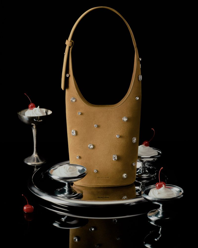 Tan handbag with crystal embellishments on a mirrored silver platter with dessert cups and cherries against a black backdrop.