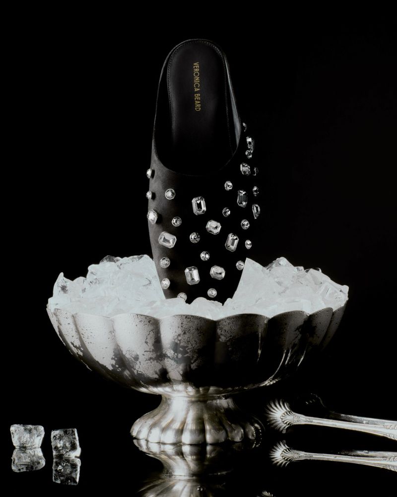 Elegant black leather mule shoe with sparkling gemstone details displayed in a silver ice bucket against a solid black backdrop.