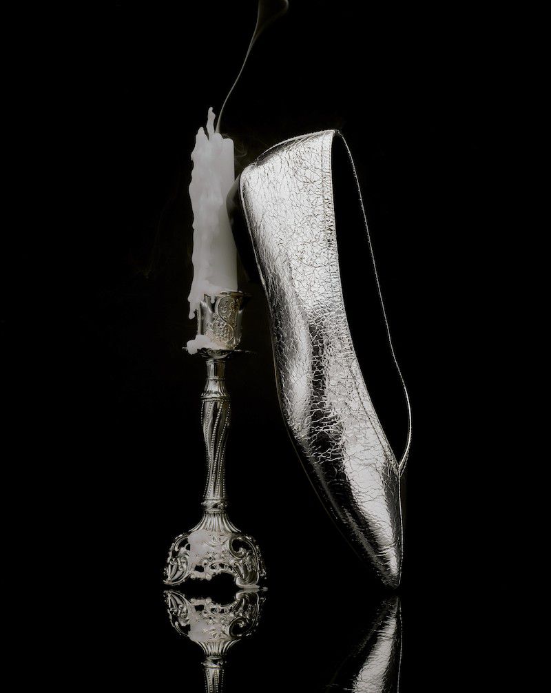 Silver high heel shoe is propped against a lit candle on a silver candlestick with a black background and reflective surface.