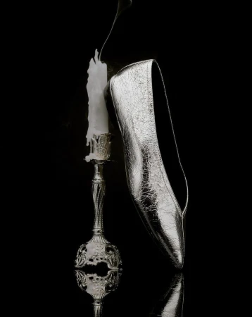 Silver high heel shoe is propped against a lit candle on a silver candlestick with a black background and reflective surface.