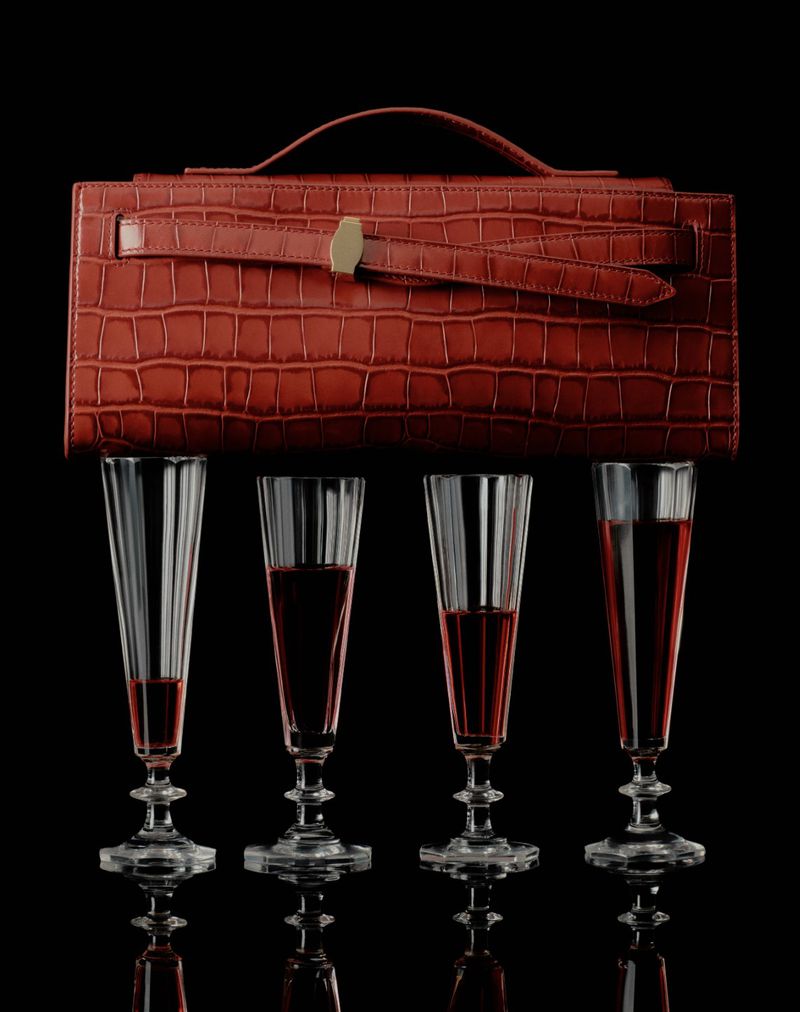 A red crocodile-skin purse rests above four champagne flutes filled with red wine, set against a stark black background.