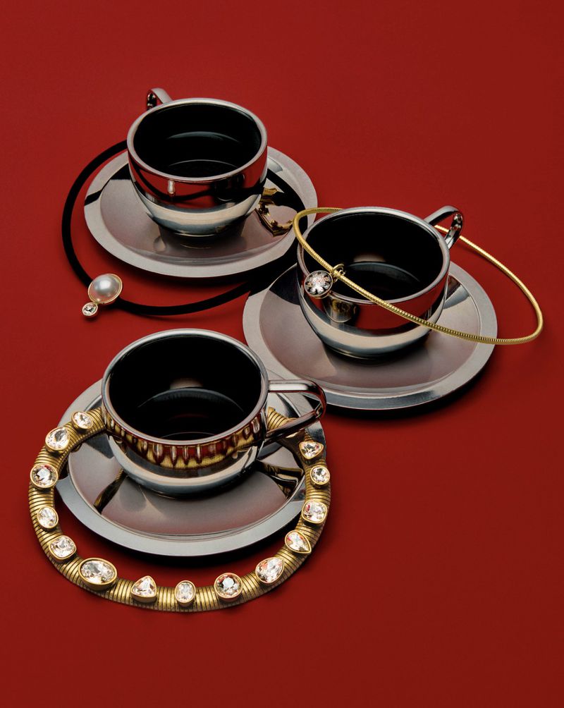 Three silver teacups on saucers are arranged on a red surface, adorned with gold and jeweled bracelets and a black hair tie.