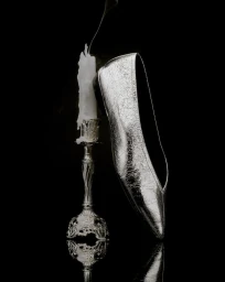 Metallic silver pointed flat shoe resting against an ornate silver candlestick with a melting white candle on a black reflective surface.