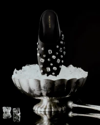 Crystal-embellished black mule set upright in a fluted silver champagne bowl filled with ice on a reflective black background.