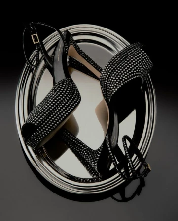 A pair of black crystal-embellished high-heeled sandals arranged on a reflective silver oval tray against a black background.