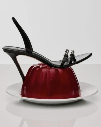 A black patent slingback stiletto sandal balanced atop a glossy red molded gelatin dessert on a white plate in a studio setting.