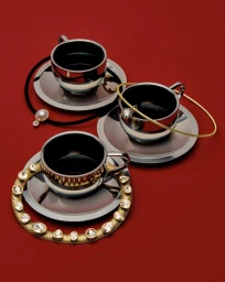 Three chrome espresso cups on matching saucers adorned with gold and crystal jewellery against a solid red surface.