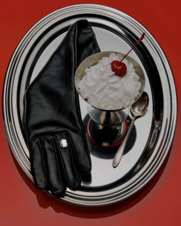 A black leather glove with a gemstone ring resting beside a whipped cream dessert with a cherry on a silver platter over a red backdrop.
