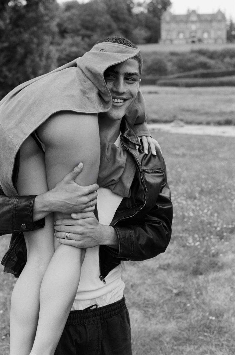 A smiling man in a black jacket carries a person draped in a light-colored garment in a grassy field with a building in the background.