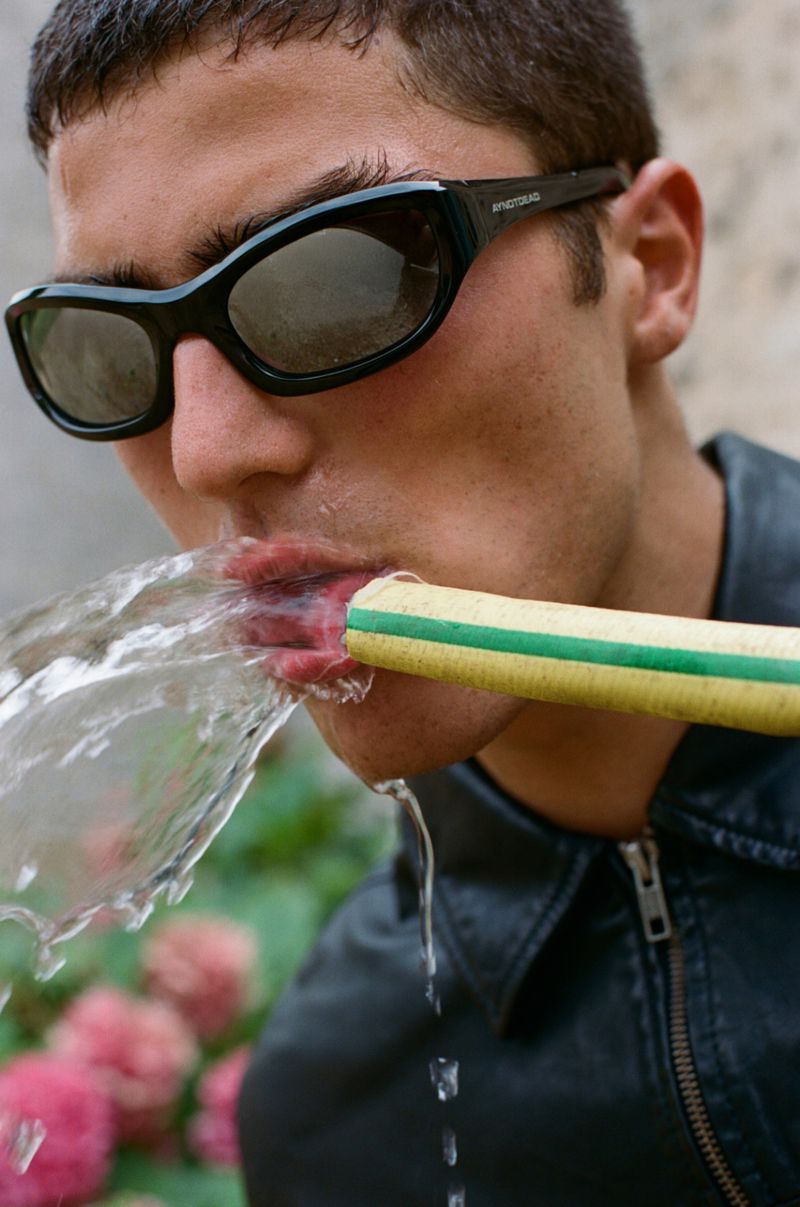 Adult man drinking from a yellow garden hose outdoors, wearing sunglasses and a black leather jacket against a floral background.