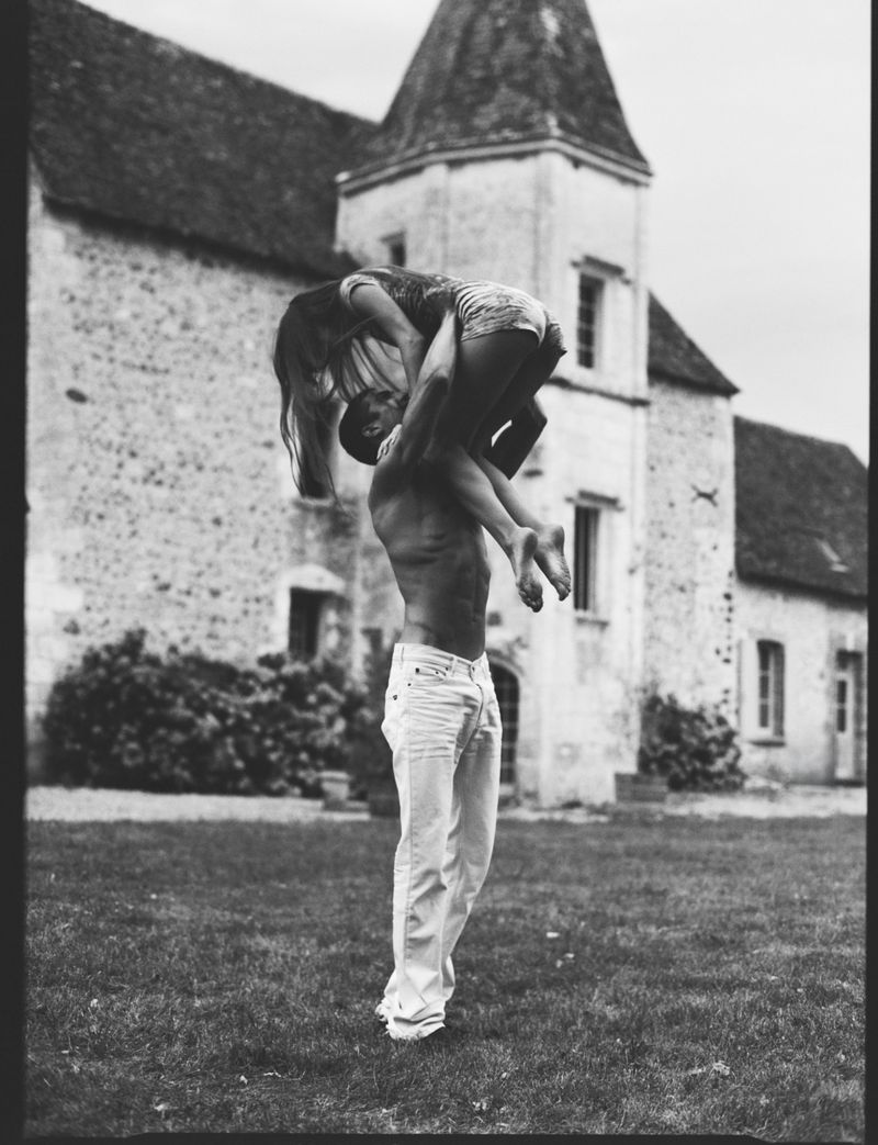 Monochrome image of a man lifting a woman in front of a stone building and grassy lawn, creating a romantic and joyful scene.