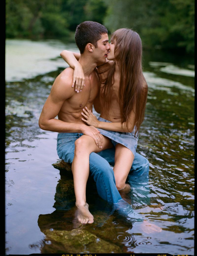 A shirtless man and woman are kissing while seated in a shallow river, embracing each other with a lush green forest backdrop.