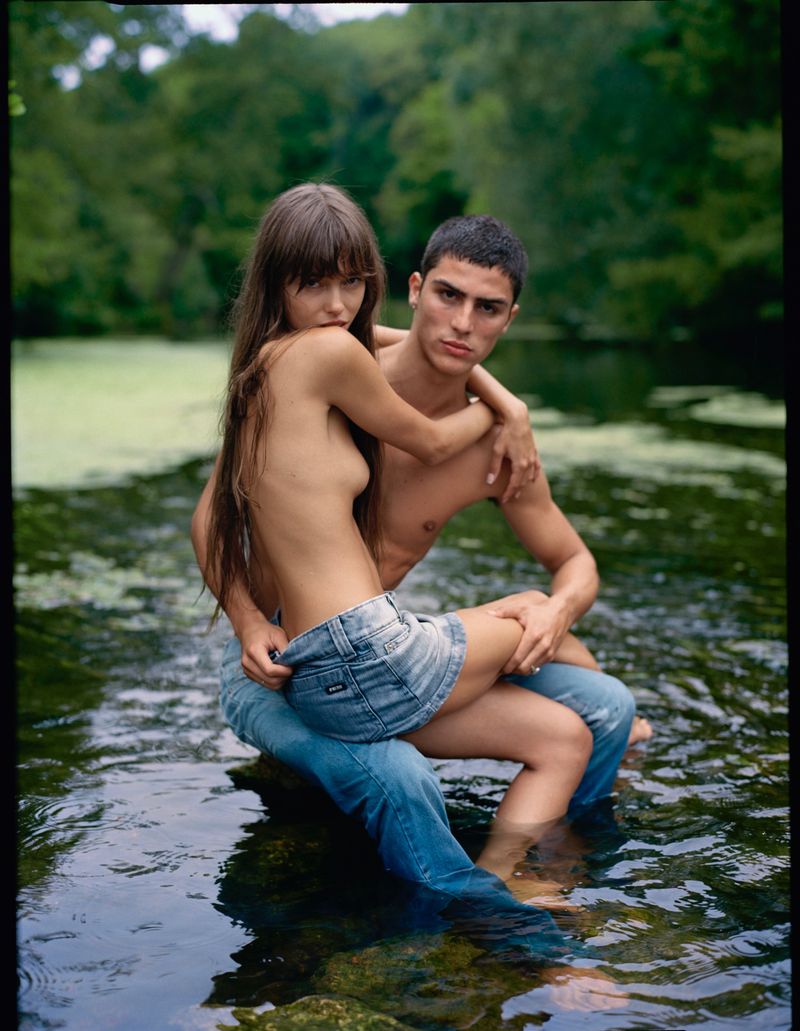 A couple embraces while sitting together in a shallow river, with natural green foliage and trees visible in the background setting.