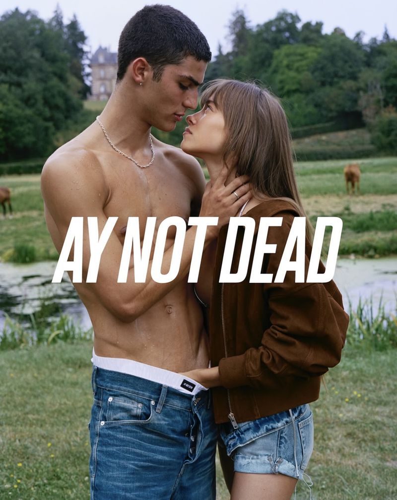 A young couple embraces outdoors in a sun-drenched field; the man is shirtless, the woman wears a brown jacket and denim shorts.